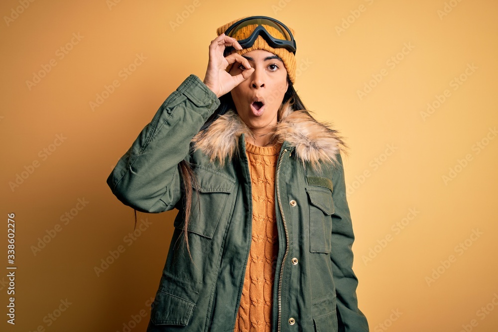 Young beautiful hispanic woman wearing ski glasses and coat for winter weather doing ok gesture shocked with surprised face, eye looking through fingers. Unbelieving expression.