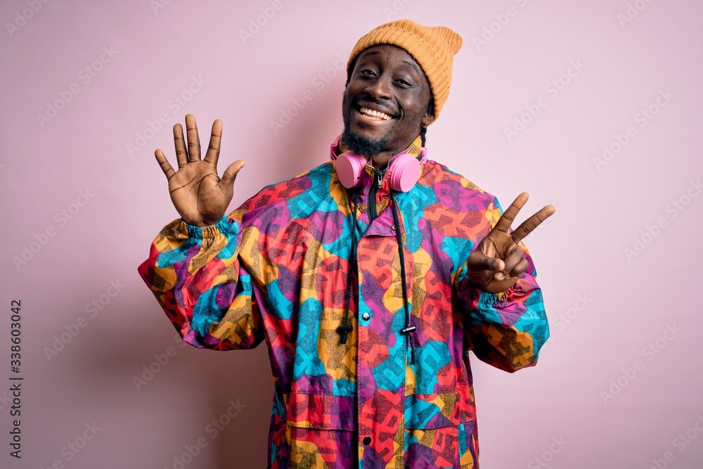 Young handsome african american man wearing colorful coat and cap over pink background showing and pointing up with fingers number seven while smiling confident and happy.