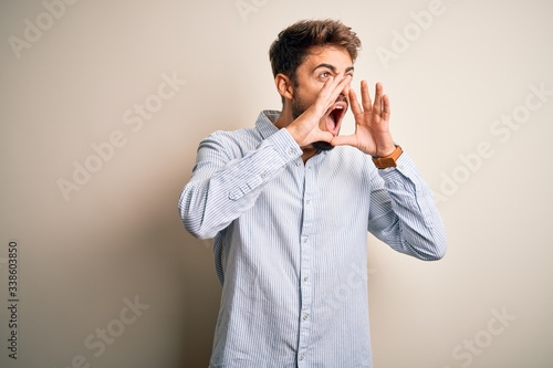 Young handsome man with beard wearing striped shirt standing over white background Shouting angry out loud with hands over mouth