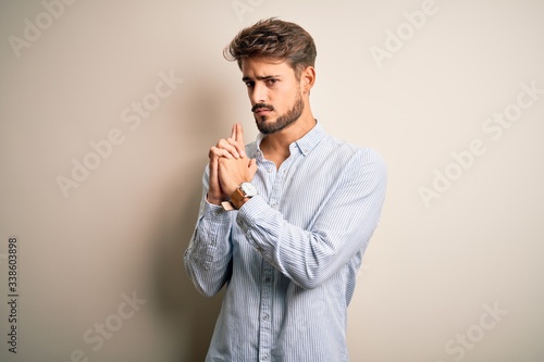 Young handsome man with beard wearing striped shirt standing over white background Holding symbolic gun with hand gesture, playing killing shooting weapons, angry face © Krakenimages.com