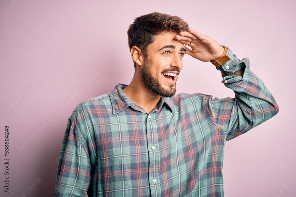Young handsome man with beard wearing casual shirt standing over pink background very happy and smiling looking far away with hand over head. Searching concept. Young handsome man with beard wearing casual shirt standing over pink background very happy and smiling looking far away with hand over head. Searching concept.