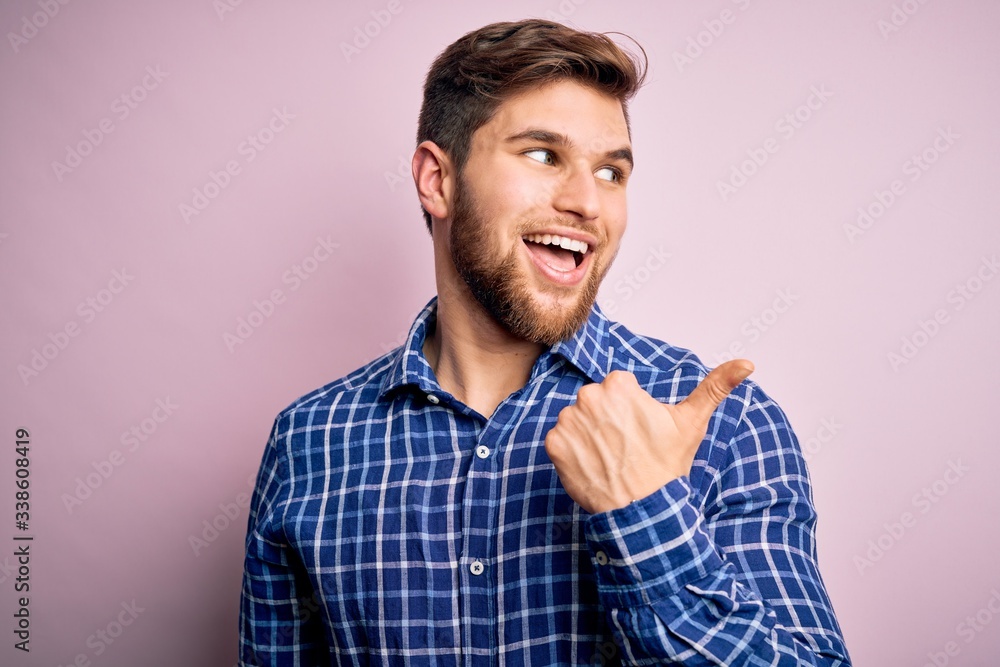 Young handsome blond man with beard and blue eyes wearing casual shirt standing smiling with happy face looking and pointing to the side with thumb up. Young handsome blond man with beard and blue eyes wearing casual shirt standing smiling with happy face looking and pointing to the side with thumb up.