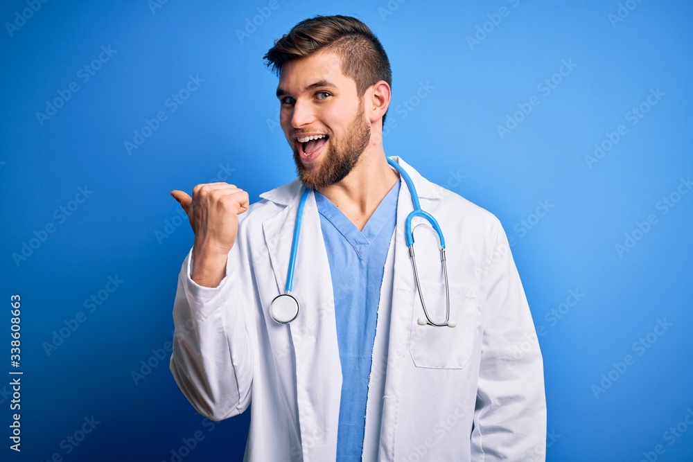 Young blond doctor man with beard and blue eyes wearing white coat and stethoscope smiling with happy face looking and pointing to the side with thumb up. Young blond doctor man with beard and blue eyes wearing white coat and stethoscope smiling with happy face looking and pointing to the side with thumb up.