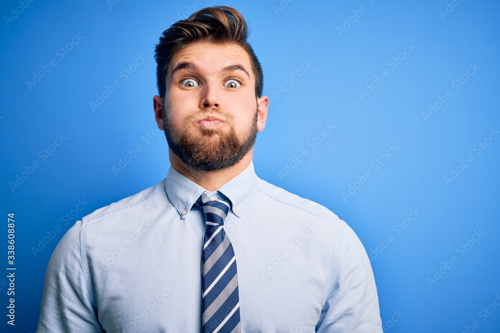 Young blond businessman with beard and blue eyes wearing elegant shirt and tie standing puffing cheeks with funny face. Mouth inflated with air, crazy expression. Young blond businessman with beard and blue eyes wearing elegant shirt and tie standing puffing cheeks with funny face. Mouth inflated with air, crazy expression.