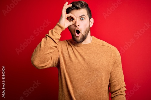 Young blond man with beard and blue eyes wearing casual sweater over red background doing ok gesture shocked with surprised face, eye looking through fingers. Unbelieving expression.