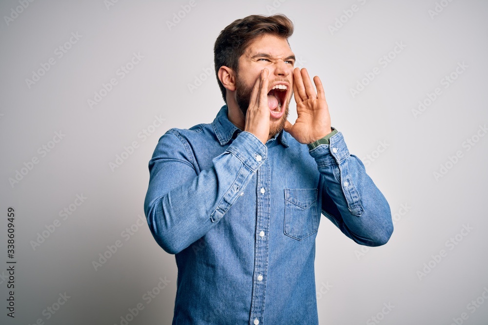 Young handsome blond man with beard and blue eyes wearing casual denim shirt Shouting angry out loud with hands over mouth Young handsome blond man with beard and blue eyes wearing casual denim shirt Shouting angry out loud with hands over mouth