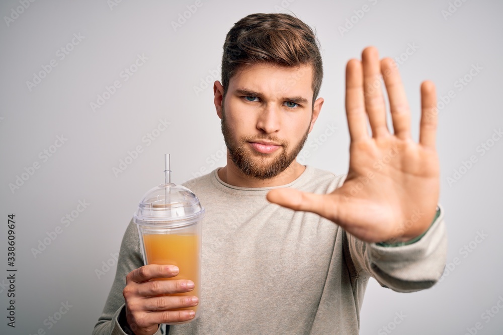 Young blond man with beard and blue eyes drinking healthy orange smoothie with open hand doing stop sign with serious and confident expression, defense gesture Young blond man with beard and blue eyes drinking healthy orange smoothie with open hand doing stop sign with serious and confident expression, defense gesture