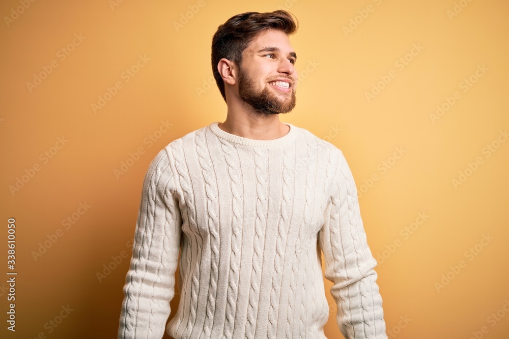Young blond man with beard and blue eyes wearing white sweater over yellow background looking away to side with smile on face, natural expression. Laughing confident. Young blond man with beard and blue eyes wearing white sweater over yellow background looking away to side with smile on face, natural expression. Laughing confident.
