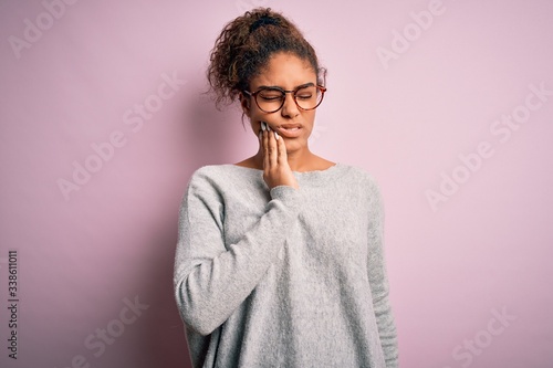 Young beautiful african american girl wearing sweater and glasses over pink background touching mouth with hand with painful expression because of toothache or dental illness on teeth. Dentist