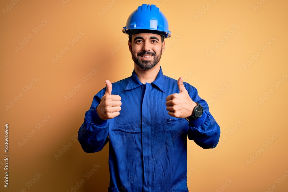 Mechanic man with beard wearing blue uniform and safety helmet over yellow background success sign doing positive gesture with hand, thumbs up smiling and happy. Cheerful expression and winner