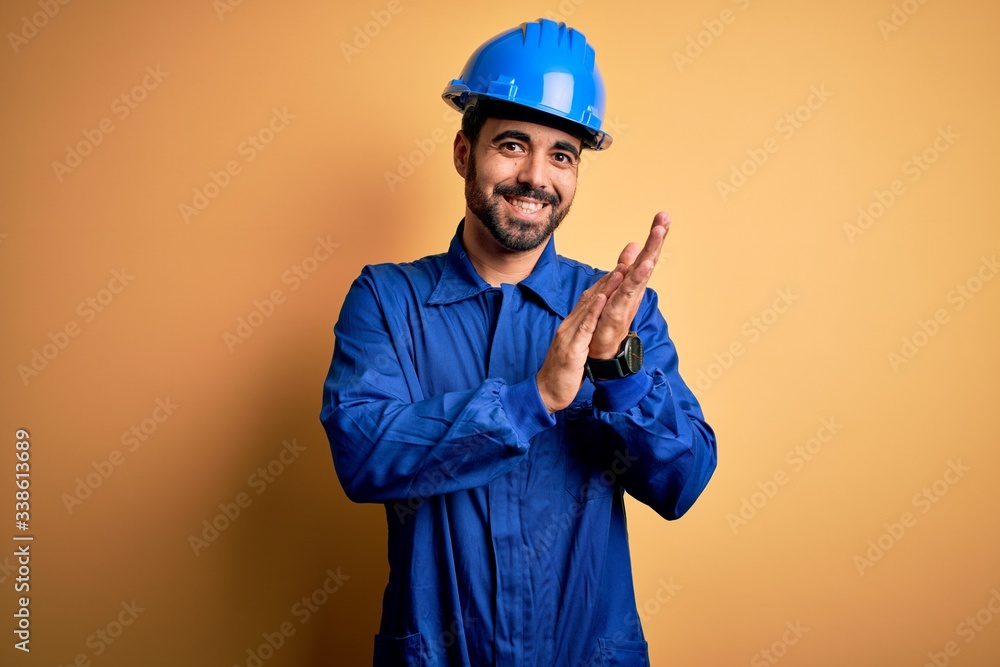 Mechanic man with beard wearing blue uniform and safety helmet over yellow background clapping and applauding happy and joyful, smiling proud hands together