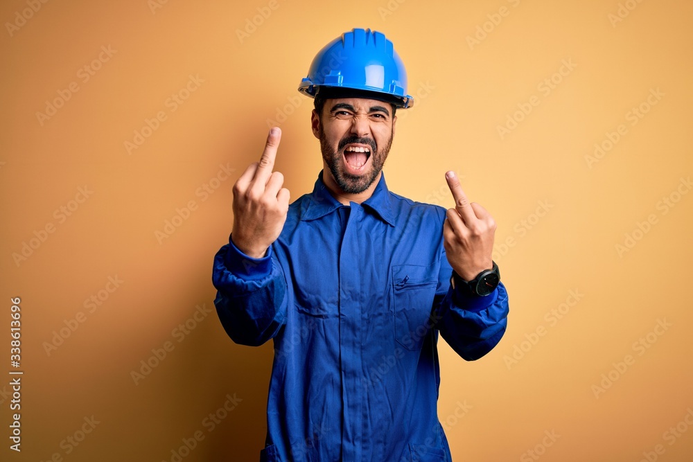 Mechanic man with beard wearing blue uniform and safety helmet over yellow background Showing middle finger doing fuck you bad expression, provocation and rude attitude. Screaming excited