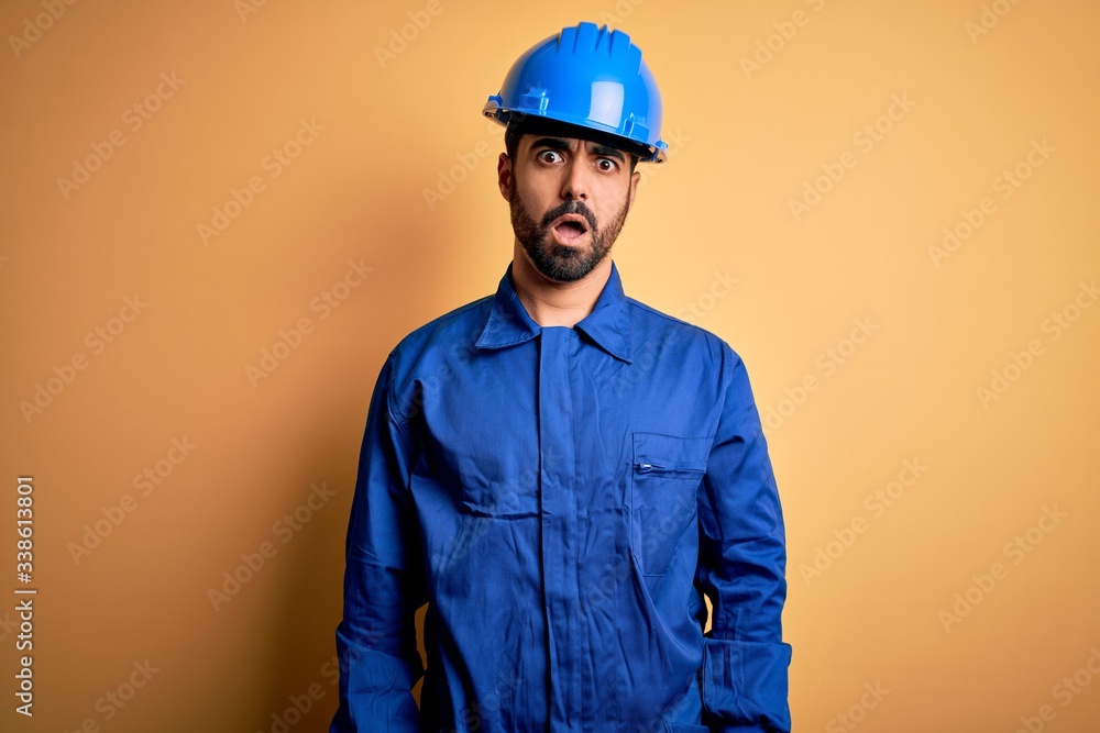 Mechanic man with beard wearing blue uniform and safety helmet over yellow background In shock face, looking skeptical and sarcastic, surprised with open mouth