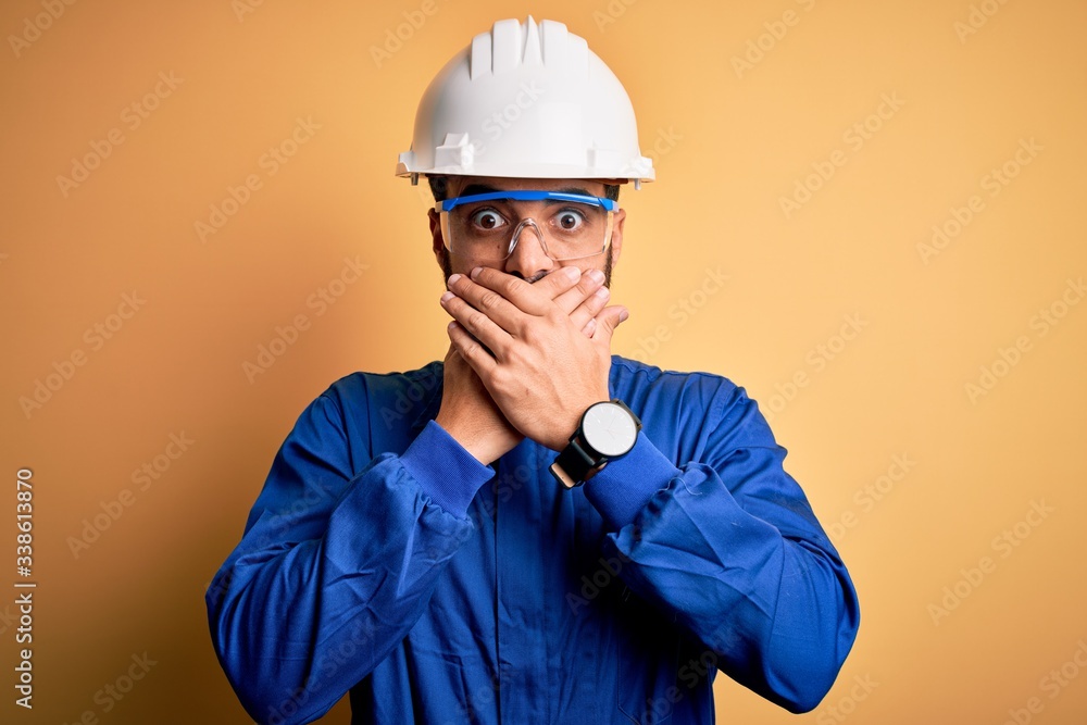 Mechanic man with beard wearing blue uniform and safety glasses over yellow background shocked covering mouth with hands for mistake. Secret concept.
