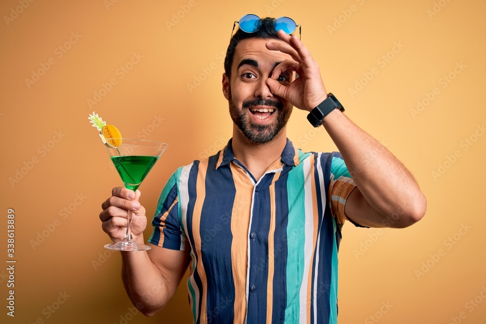 Young handsome tourist man with beard on vacation wearing summer shirt drinking cocktail with happy face smiling doing ok sign with hand on eye looking through fingers Young handsome tourist man with beard on vacation wearing summer shirt drinking cocktail with happy face smiling doing ok sign with hand on eye looking through fingers