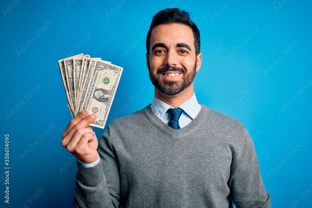 Young handsome man with beard holding bunch of dollars banknotes over blue background with a happy face standing and smiling with a confident smile showing teeth Young handsome man with beard holding bunch of dollars banknotes over blue background with a happy face standing and smiling with a confident smile showing teeth