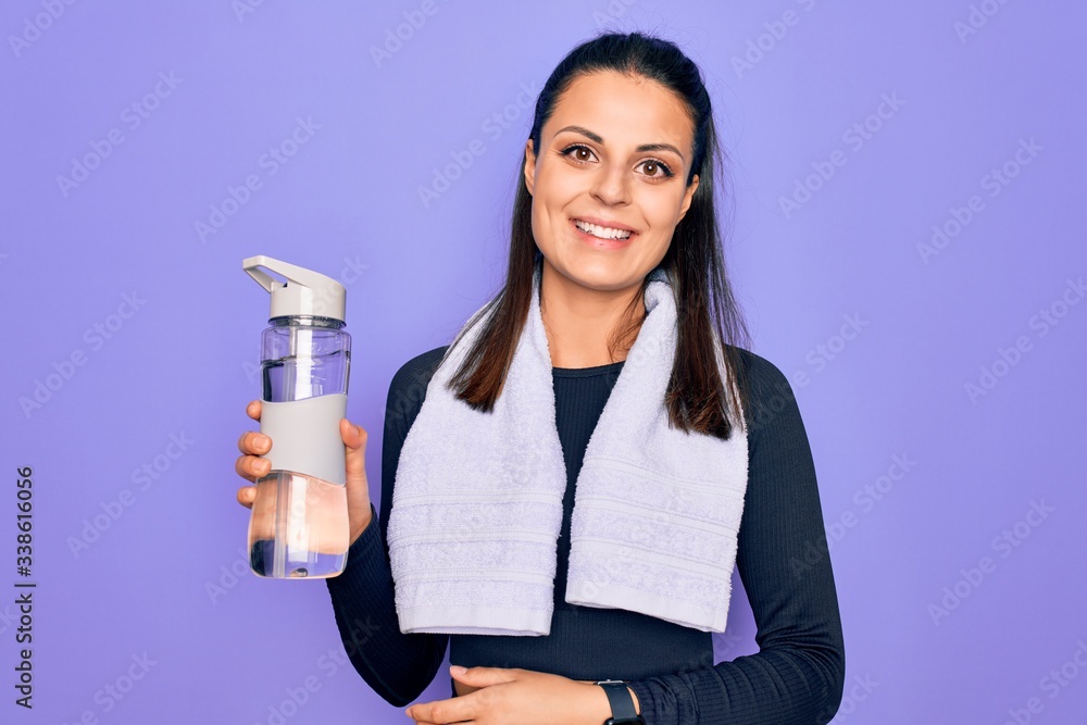 Young beautiful brunette sporty woman using sport towel drinking bottle of water looking positive and happy standing and smiling with a confident smile showing teeth Young beautiful brunette sporty woman using sport towel drinking bottle of water looking positive and happy standing and smiling with a confident smile showing teeth