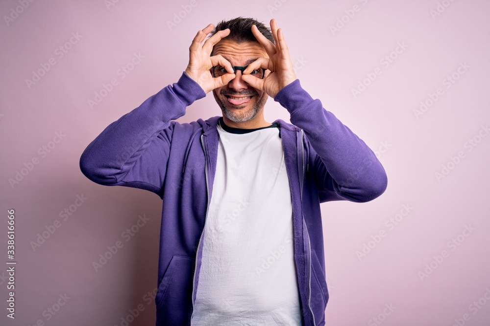 Young handsome man wearing purple sweatshirt and glasses standing over pink background doing ok gesture like binoculars sticking tongue out, eyes looking through fingers. Crazy expression. Young handsome man wearing purple sweatshirt and glasses standing over pink background doing ok gesture like binoculars sticking tongue out, eyes looking through fingers. Crazy expression.