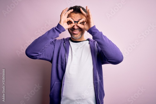 Young handsome man wearing purple sweatshirt and glasses standing over pink background doing ok gesture like binoculars sticking tongue out, eyes looking through fingers. Crazy expression. © Krakenimages.com
