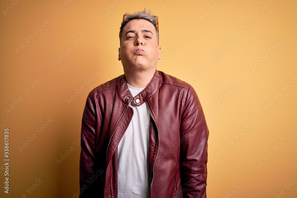 Young handsome modern man wearing fashion leather jacket and sunglasses over yellow background puffing cheeks with funny face. Mouth inflated with air, crazy expression. Young handsome modern man wearing fashion leather jacket and sunglasses over yellow background puffing cheeks with funny face. Mouth inflated with air, crazy expression.