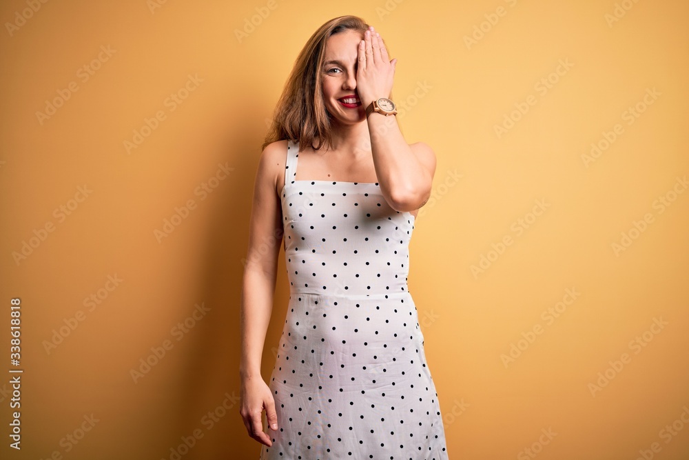 Young beautiful blonde woman on vacation wearing summer dress over yellow background covering one eye with hand, confident smile on face and surprise emotion. Young beautiful blonde woman on vacation wearing summer dress over yellow background covering one eye with hand, confident smile on face and surprise emotion.