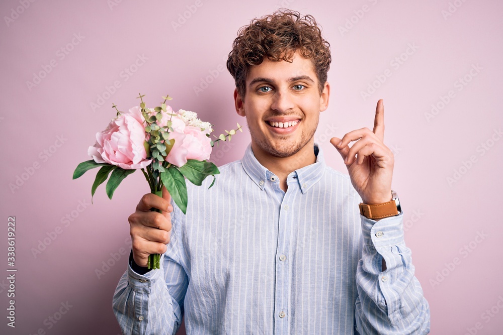 Young blond man with curly hair holding beautiful bouquet over isolated pink background surprised with an idea or question pointing finger with happy face, number one Young blond man with curly hair holding beautiful bouquet over isolated pink background surprised with an idea or question pointing finger with happy face, number one