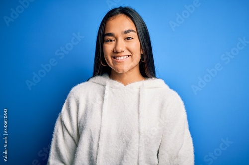 Young beautiful asian sportswoman wearing sweatshirt standing over isolated blue background with a happy and cool smile on face. Lucky person.