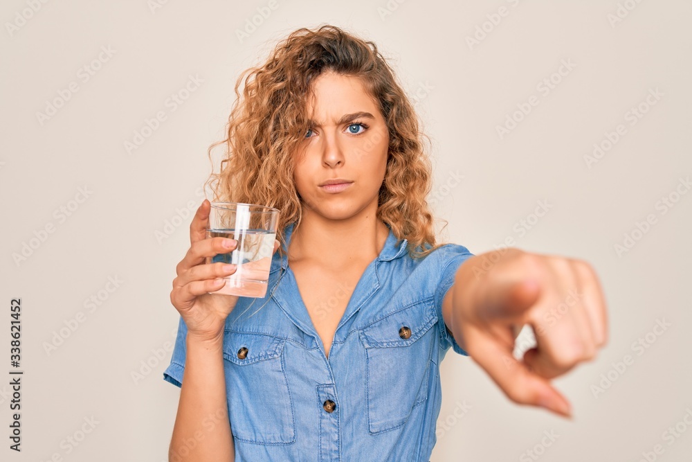Young beautiful blonde woman with blue eyes drinking glass of water over white background pointing with finger to the camera and to you, hand sign, positive and confident gesture from the front Young beautiful blonde woman with blue eyes drinking glass of water over white background pointing with finger to the camera and to you, hand sign, positive and confident gesture from the front
