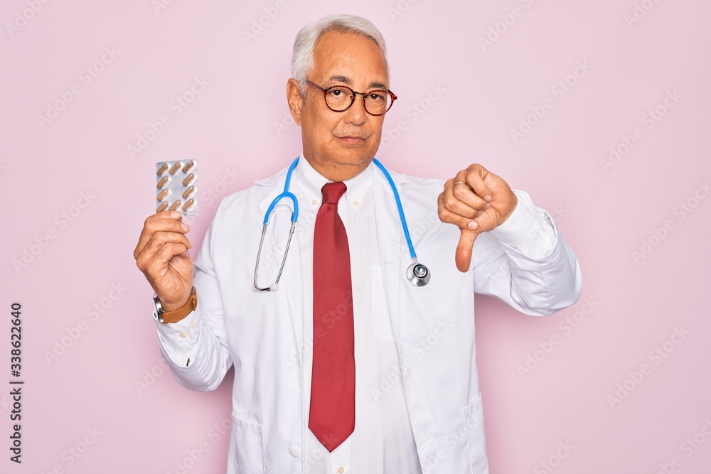 Middle age senior grey-haired doctor man holding prescription pills over pink background with angry face, negative sign showing dislike with thumbs down, rejection concept Middle age senior grey-haired doctor man holding prescription pills over pink background with angry face, negative sign showing dislike with thumbs down, rejection concept