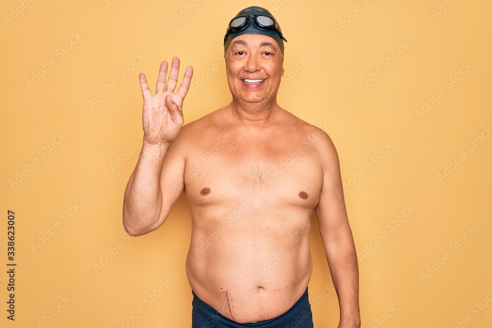 Middle age senior grey-haired swimmer man wearing swimsuit, cap and goggles showing and pointing up with fingers number four while smiling confident and happy.