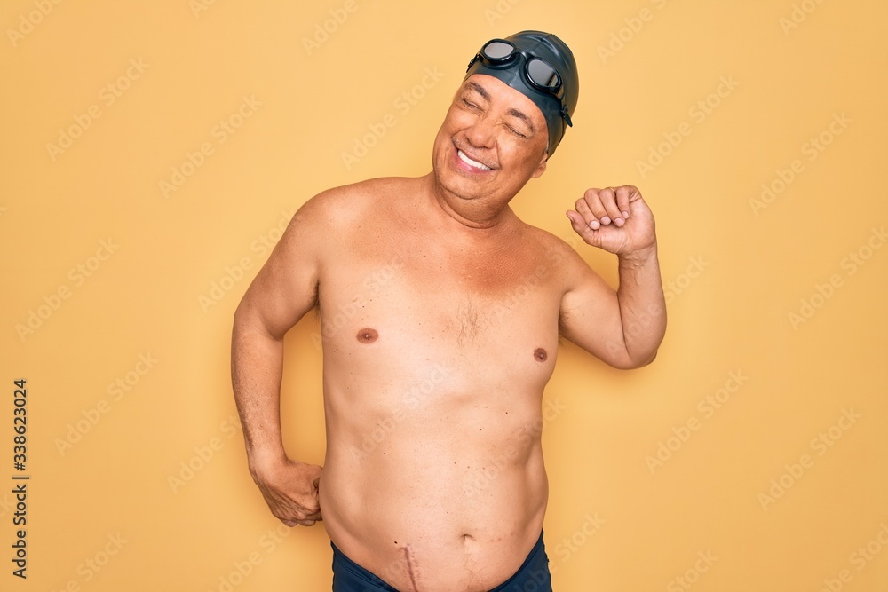 Middle age senior grey-haired swimmer man wearing swimsuit, cap and goggles stretching back, tired and relaxed, sleepy and yawning for early morning