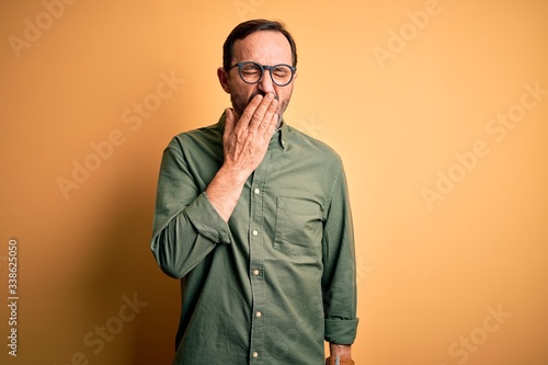 Middle age hoary man wearing casual green shirt and glasses over isolated yellow background bored yawning tired covering mouth with hand. Restless and sleepiness.