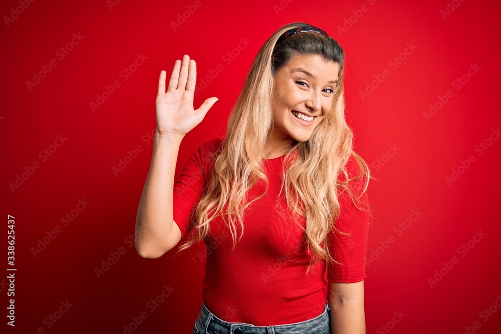 Young beautiful blonde woman wearing casual t-shirt standing over isolated red background Waiving saying hello happy and smiling, friendly welcome gesture Young beautiful blonde woman wearing casual t-shirt standing over isolated red background Waiving saying hello happy and smiling, friendly welcome gesture