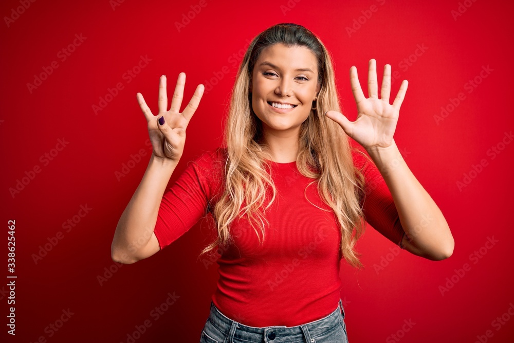 Young beautiful blonde woman wearing casual t-shirt standing over isolated red background showing and pointing up with fingers number nine while smiling confident and happy. Young beautiful blonde woman wearing casual t-shirt standing over isolated red background showing and pointing up with fingers number nine while smiling confident and happy.
