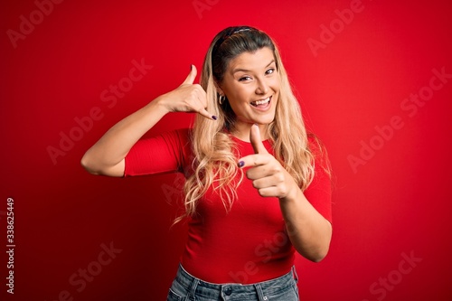Young beautiful blonde woman wearing casual t-shirt standing over isolated red background smiling doing talking on the telephone gesture and pointing to you. Call me.
