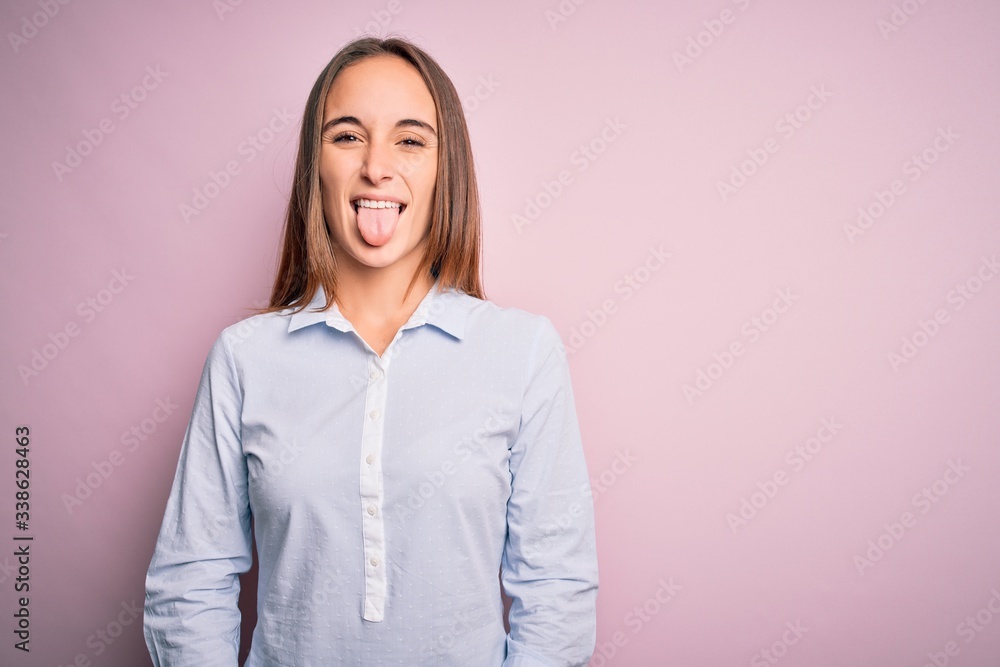 Young beautiful businesswoman wearing elegant shirt standing over isolated pink background sticking tongue out happy with funny expression. Emotion concept. Young beautiful businesswoman wearing elegant shirt standing over isolated pink background sticking tongue out happy with funny expression. Emotion concept.