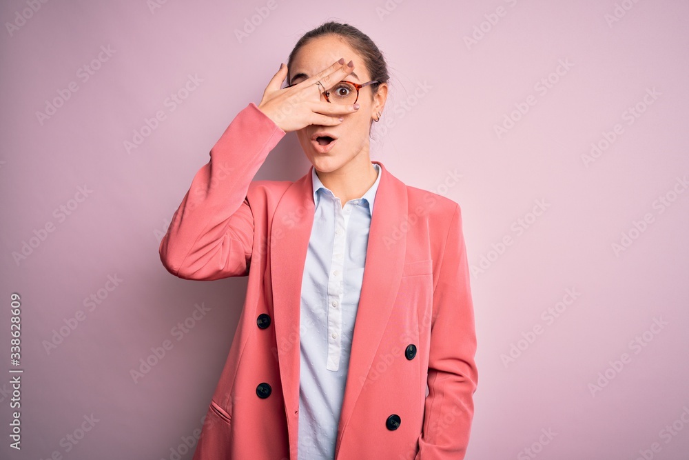 Young beautiful businesswoman wearing jacket and glasses over isolated pink background peeking in shock covering face and eyes with hand, looking through fingers with embarrassed expression. Young beautiful businesswoman wearing jacket and glasses over isolated pink background peeking in shock covering face and eyes with hand, looking through fingers with embarrassed expression.