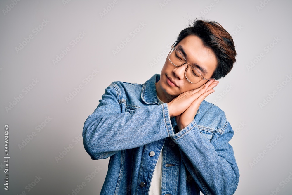 Young handsome chinese man wearing denim jacket and glasses over white background sleeping tired dreaming and posing with hands together while smiling with closed eyes. Young handsome chinese man wearing denim jacket and glasses over white background sleeping tired dreaming and posing with hands together while smiling with closed eyes.