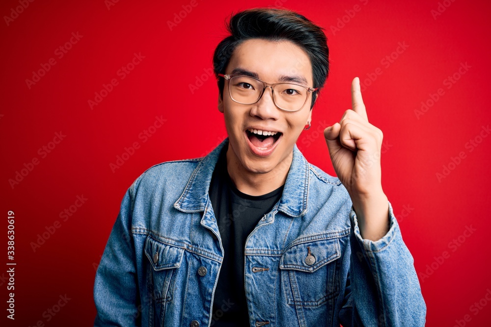 Young handsome chinese man wearing denim jacket and glasses over red background pointing finger up with successful idea. Exited and happy. Number one. Young handsome chinese man wearing denim jacket and glasses over red background pointing finger up with successful idea. Exited and happy. Number one.