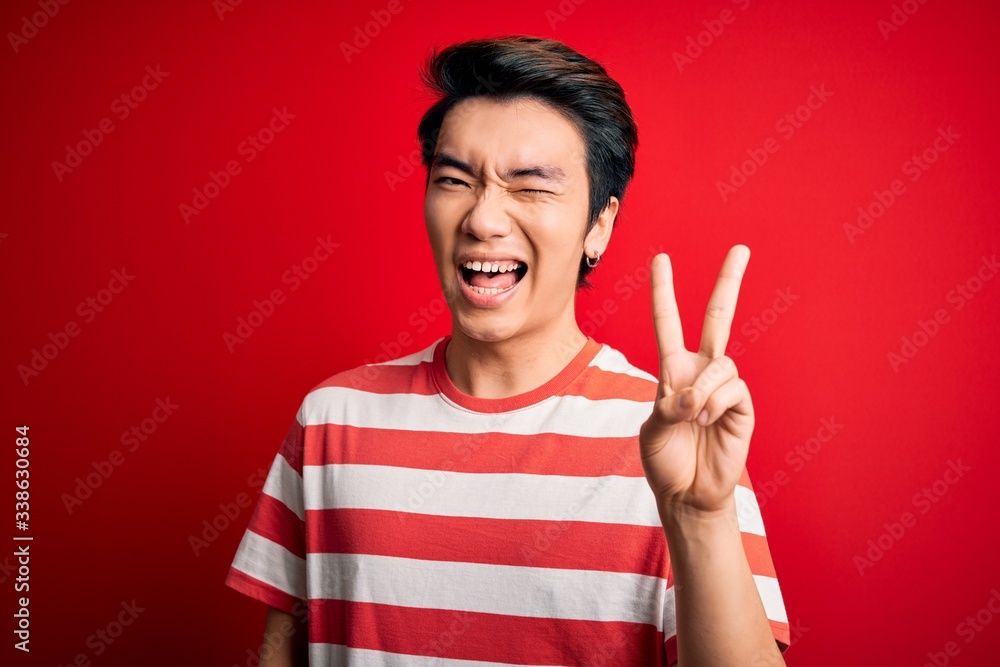 Young handsome chinese man wearing casual striped t-shirt standing over red background smiling with happy face winking at the camera doing victory sign with fingers. Number two. Young handsome chinese man wearing casual striped t-shirt standing over red background smiling with happy face winking at the camera doing victory sign with fingers. Number two.