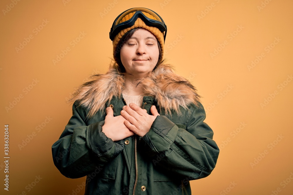 Young down syndrome woman wearing ski coat and glasses for winter weather smiling with hands on chest with closed eyes and grateful gesture on face. Health concept.