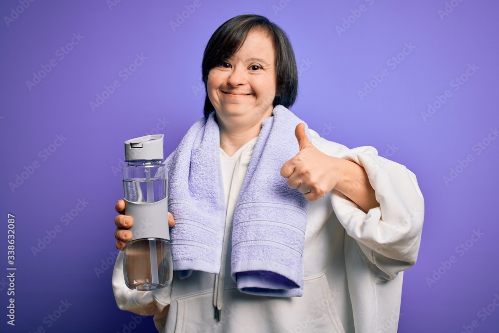 Young down syndrome fitness woman training at gym holding water bottle and sport towel happy with big smile doing ok sign, thumb up with fingers, excellent sign Young down syndrome fitness woman training at gym holding water bottle and sport towel happy with big smile doing ok sign, thumb up with fingers, excellent sign