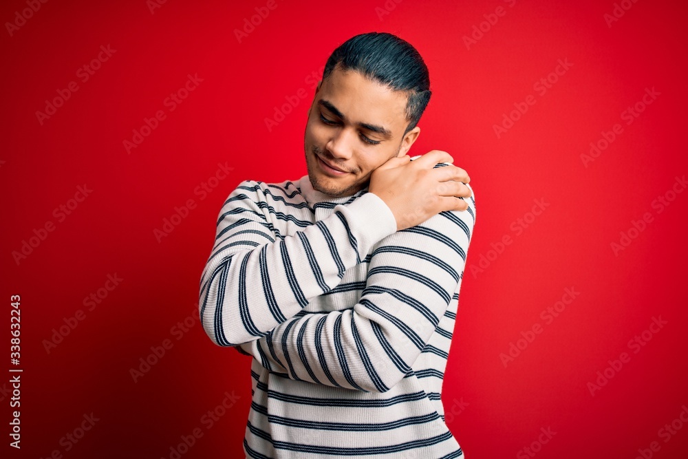 Young brazilian man wearing casual striped t-shirt standing over isolated red background Hugging oneself happy and positive, smiling confident. Self love and self care Young brazilian man wearing casual striped t-shirt standing over isolated red background Hugging oneself happy and positive, smiling confident. Self love and self care
