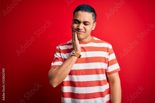 Young brazilian man wearing casual striped t-shirt standing over isolated red background touching mouth with hand with painful expression because of toothache or dental illness on teeth. Dentist © Krakenimages.com