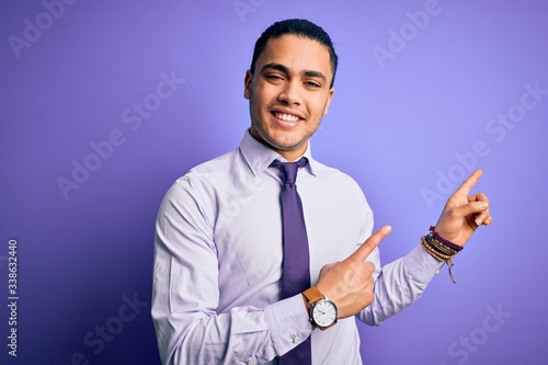 Young brazilian businessman wearing elegant tie standing over isolated purple background smiling and looking at the camera pointing with two hands and fingers to the side.