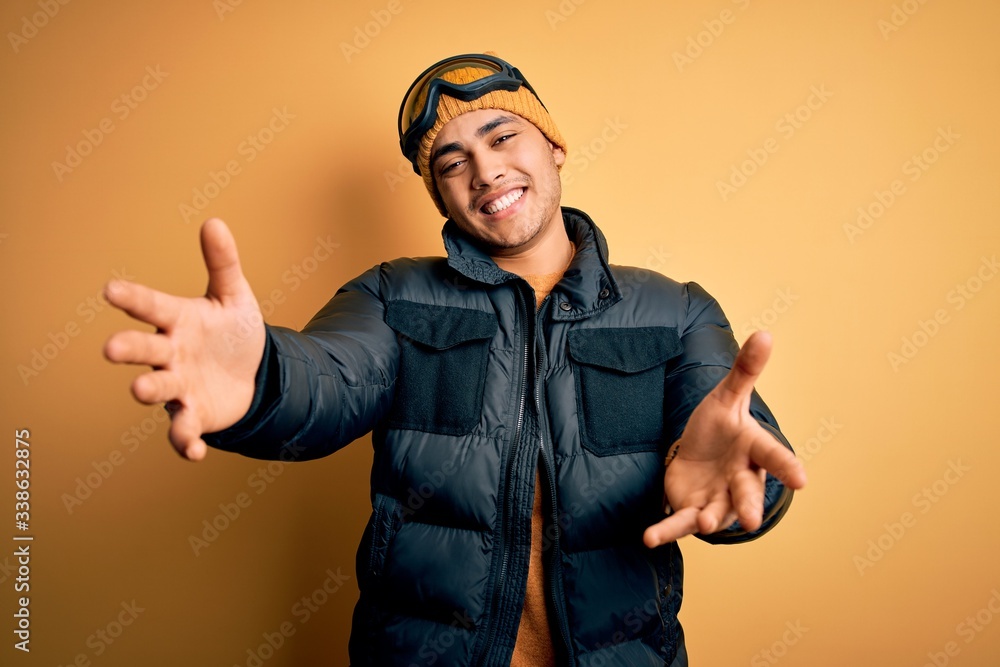 Young brazilian skier man wearing snow sportswear and ski goggles over yellow background looking at the camera smiling with open arms for hug. Cheerful expression embracing happiness. Young brazilian skier man wearing snow sportswear and ski goggles over yellow background looking at the camera smiling with open arms for hug. Cheerful expression embracing happiness.
