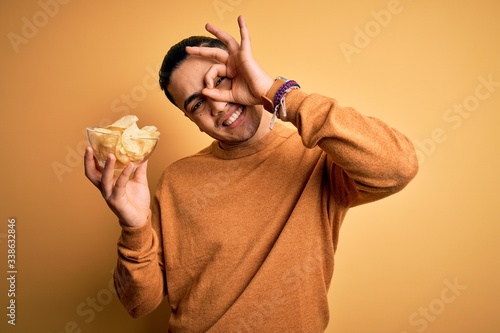 Young brazilian man holding bowl with chips potatoes over isolated yellow background with happy face smiling doing ok sign with hand on eye looking through fingers