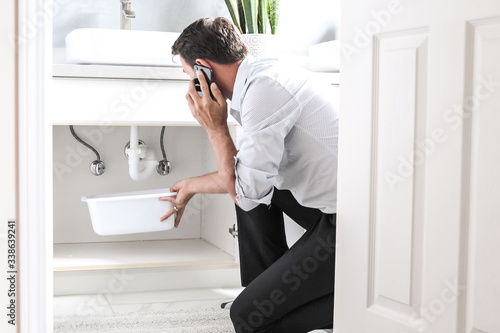 Man Calling Plumber In Front Of Water Leaking From Sink Pipe