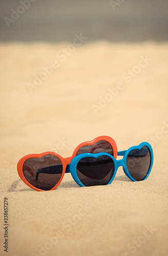 Blue and red sunglasses in shape of heart on sand at beach. Summer time concept