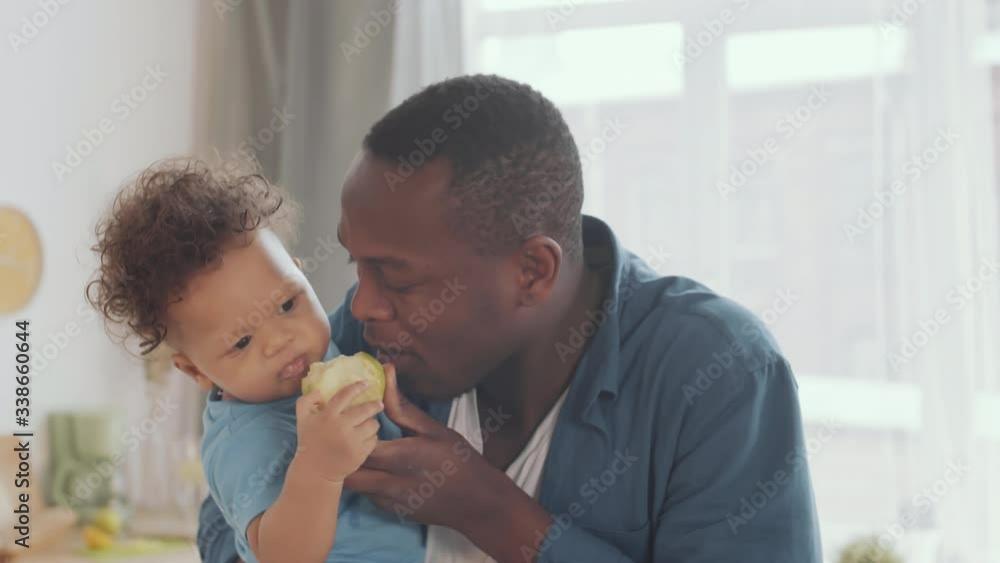 Medium shot of young attractive African man holding cute kid on his hands and showing him how to bite and eat apple Medium shot of young attractive African man holding cute kid on his hands and showing him how to bite and eat apple